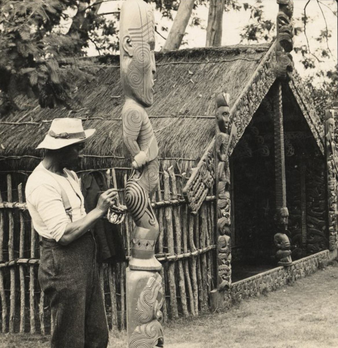 Egy maori férfi retusálja a tetoválást egy tiki szoboron.